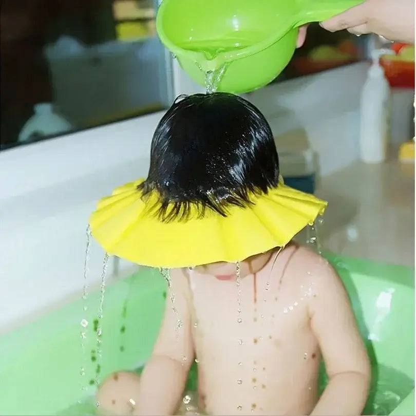 A child in a green bath, wearing a yellow shampoo shield while water is poured over their head.
