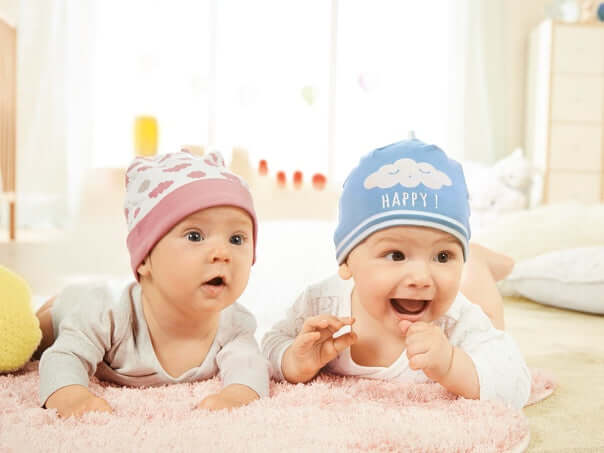 Two happy babies in colorful beanies, playing on a soft rug in a bright, cozy room.