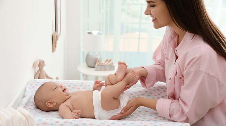Mother changing diaper of her happy infant on a changing table with soft pastel colors.
