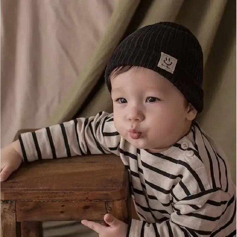 Adorable baby wearing a knitted black beanie and striped shirt, posing with a wooden chair in soft lighting.