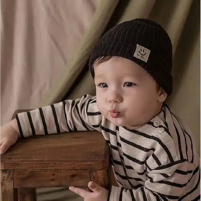 Adorable baby wearing a knitted black beanie and striped shirt, posing with a wooden chair in soft lighting.