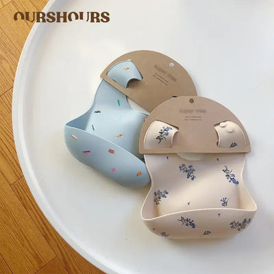 Two cute silicone baby bibs in blue and beige with playful patterns on a round tray.