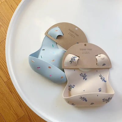 Two stylish baby bibs in soft silicone, one light blue with patterns and one cream with floral designs, displayed on a round tray.