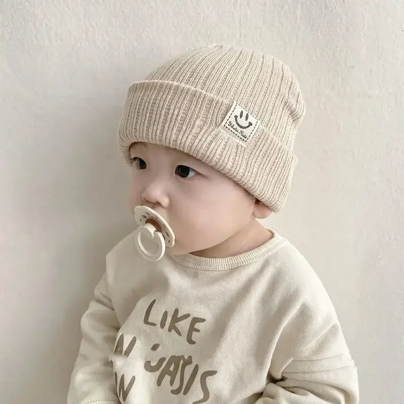 Baby wearing a beige knitted beanie hat with a smiley patch, sitting peacefully with a pacifier, dressed in a casual shirt.