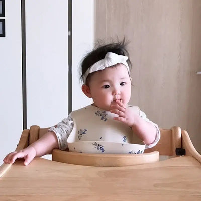 Cute toddler girl in a floral silicone bib sitting at a wooden high chair, enjoying mealtime.