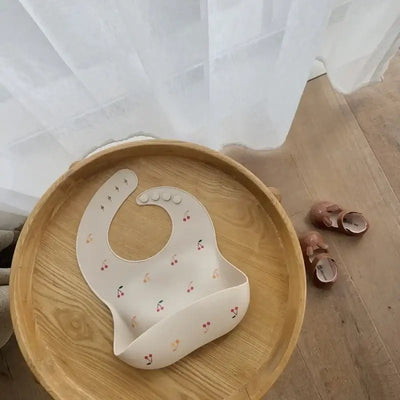 Stylish silicone baby bib with cherry pattern, placed on a wooden bowl next to cute toddler shoes.